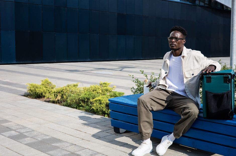African American man sitting on a blue bench with a thermal bag, enjoying a sunny day.