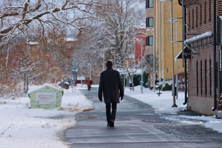 A person in a black coat walks along a snowy sidewalk in a winter cityscape.