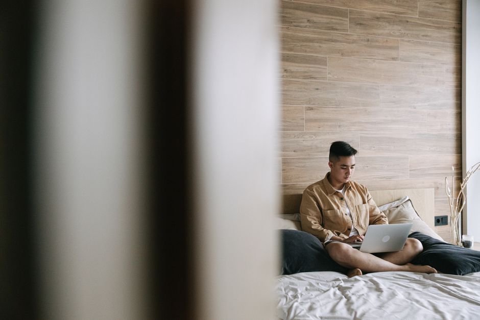 A young adult sitting on a bed using a laptop in a modern cozy bedroom with wooden decor.