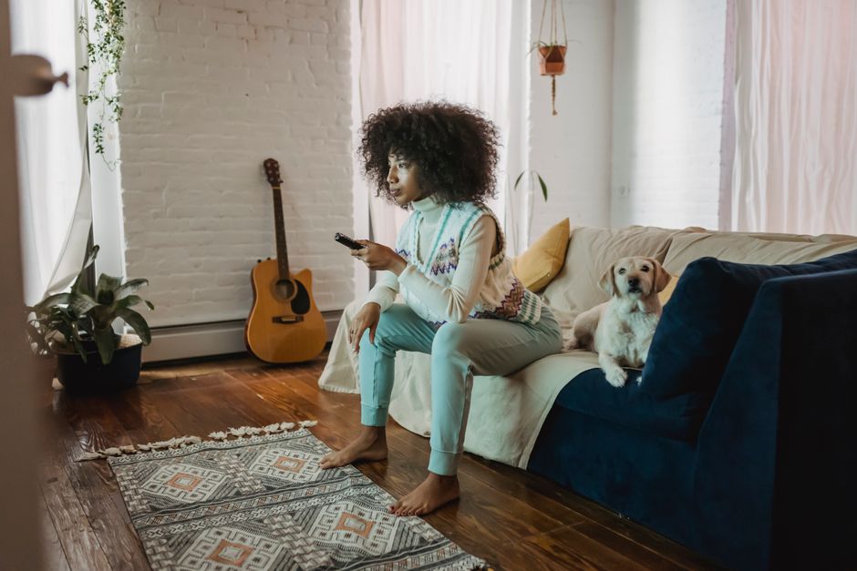 Full body of barefooted serious young African American lady with curly hair resting on sofa with obedient dog and switching TV channels with remote controller