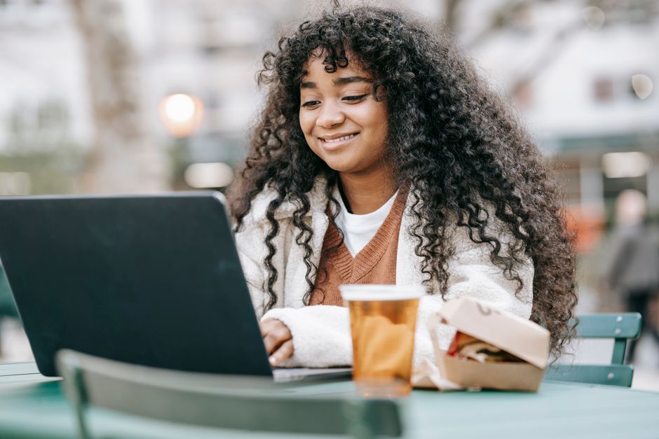 Cheerful African American female with curly hair browsing netbook while sitting at table with glass of beer and burger in outdoor restaurant