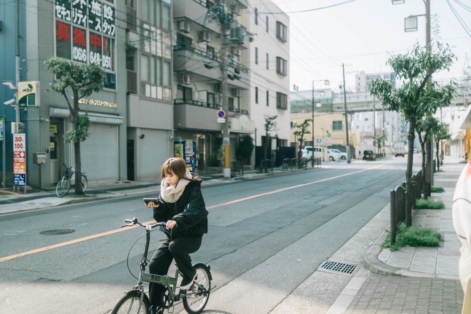 A young woman on her phone rides a bicycle through a quiet Japanese street in daylight.