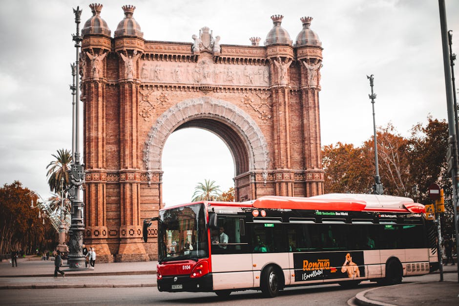 Red city bus passing by the historic Arc de Triomf in Barcelona with autumn leaves.
