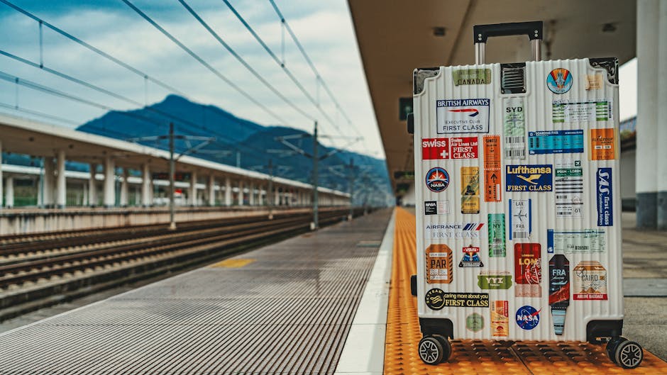 White suitcase adorned with travel stickers on empty railway platform, mountain backdrop.